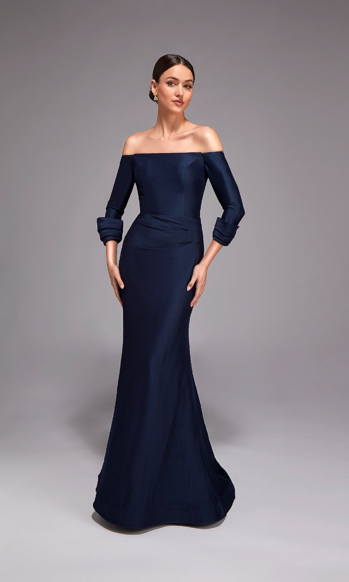 A woman stands in a studio wearing the Alyce 27782 long formal dress—navy blue, off-shoulder, fitted sleeves, stretch mikado fabric, and a mermaid silhouette with a subtle flare at the hem.